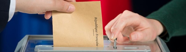 La main d'un électeur glisse une enveloppe de vote dans l'urne des élections et la main d'un élu actionne l'urne pour comptabiliser le vote